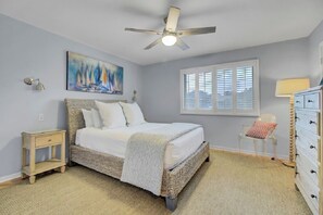 A bedroom with a bed, a ceiling fan, a window with blinds, a nightstand, a dresser, and a painting on the wall. - Award-Winning Resort Amenities | Mariners Walk 7F (Isle of Palms)