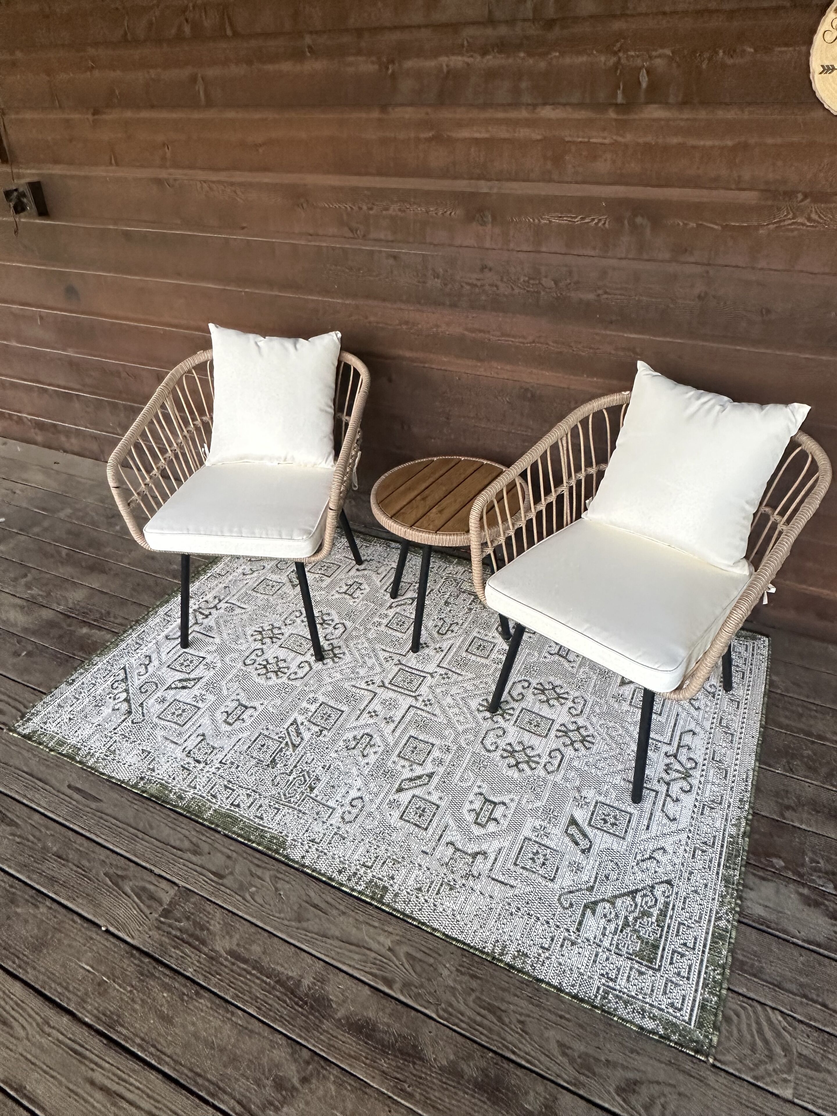 Two wicker chairs with white cushions on a patterned rug, placed on a wooden deck against a wooden wall.