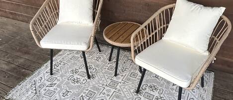 Two wicker chairs with white cushions on a patterned rug, placed on a wooden deck against a wooden wall.