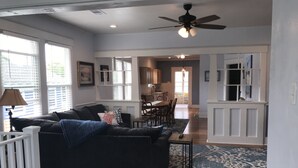 Un salón amplio con un sofá de color oscuro, una zona de comedor con muebles de madera y una cocina visible a través de una puerta abierta.