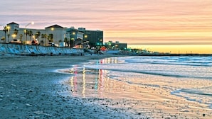 Un hotel frente a la playa con varias construcciones, una playa arenosa y una puesta de sol al fondo.