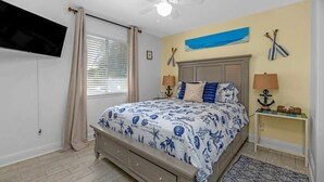 Ein Schlafzimmer mit Bett, Deckenventilator, an der Wand montiertem Fernseher, einem Fenster mit Vorhängen und einem kleinen Tisch mit Lampe.