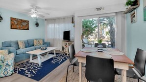 Ein Wohnzimmer mit einer Couch, einem Couchtisch und einem Esstisch mit Stühlen. Es gibt einen Fernseher, einen Deckenventilator und ein Fenster mit Blick auf Palmen.