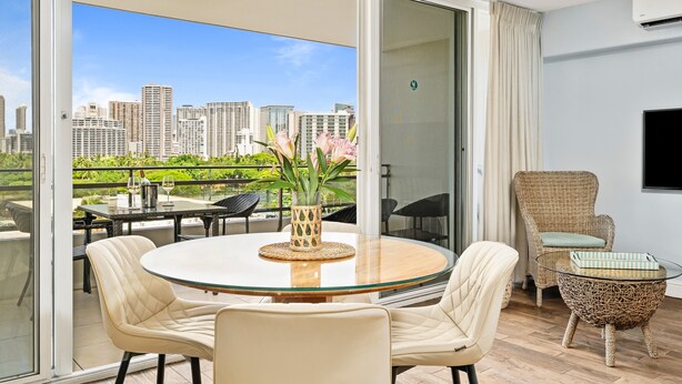 Un salon moderne avec une table en verre, des fauteuils beiges, un fauteuil en osier et une vue sur le paysage urbain, visible à travers de grandes portes vitrées.