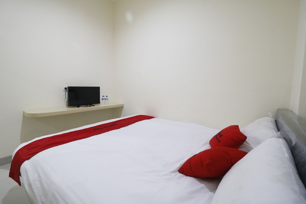 Una cama bien tendida con ropa de cama blanca y roja, un cabecero gris y una mesita de noche con un dispositivo negro.