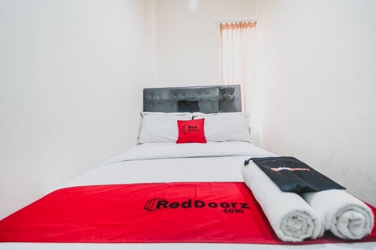 Una cama bien hecha con una colcha roja y blanca, un almohadón con etiqueta roja y toallas enrolladas sobre un tapete rojo.