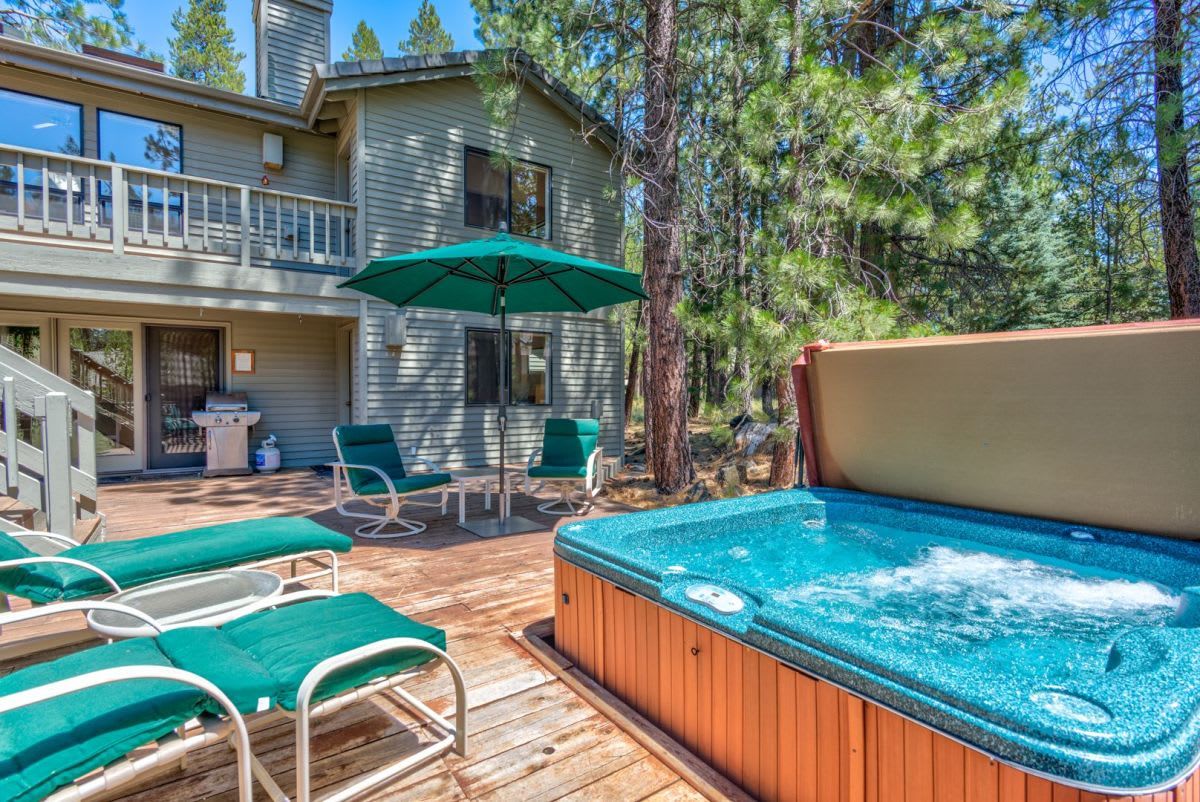 Een houten terras met loungebedden en een jacuzzi, omringd door bomen en een huis.