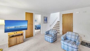 Un salon avec un téléviseur à écran plat posé sur un meuble en bois, deux fauteuils à motifs écossais, un escalier et une porte.