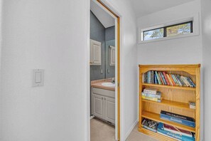A narrow hallway with a wooden bookshelf filled with books, a bathroom with a sink and mirror, and a small window above the mirror.
