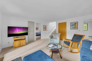 A living room with a flat-screen TV, a glass coffee table, two armchairs, a sofa, a wooden cabinet, and a staircase leading to another room.