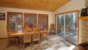 A dining area with a wooden table and chairs, large windows with blinds, a stone fireplace, and a view of a deck and trees. - Make Memories in Marla Bay, Private Homeowners Beach, 8 Guests (Zephyr Cove)