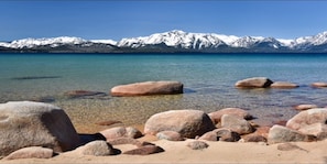 Une plage à l’eau claire, avec de gros rochers au premier plan et des montagnes enneigées en arrière-plan.