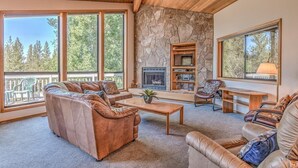 A spacious living room with leather sofas, a stone fireplace, and large windows offering a view of trees. - Panoramic mountain views hot tub, air conditioning (Sunriver)