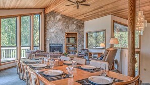 A spacious dining area with a large wooden table set for six, flanked by chairs. A stone fireplace and a television are visible in the background. - Panoramic mountain views hot tub, air conditioning (Sunriver)