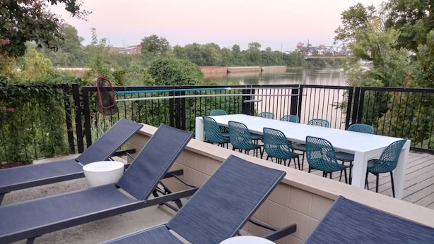 Une terrasse dotée de chaises longues et d’une table, offrant une vue sur une étendue d’eau et la verdure environnante.
