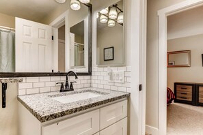 Une salle de bain avec un lavabo doté d’un plan de travail en granit, des carreaux blancs, un grand miroir et un luminaire placé au-dessus du miroir.