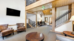 Un hall d’entrée spacieux avec un plafond haut, des poutres en bois, un escalier, des fauteuils en cuir, une table ronde en bois et un téléviseur à écran plat fixé au mur.