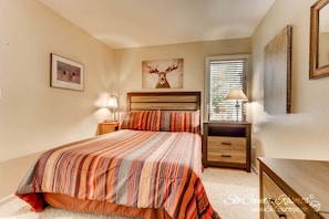 Quarto com cama grande, cabeceira de madeira, mesa de cabeceira com luminária, cômoda e janela com persianas.