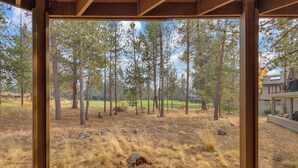 A view from a covered outdoor area with a wooden deck, looking out at a forested landscape with tall trees and a grassy field. - Unique, spacious, private deck, HT, great room (Sunriver)