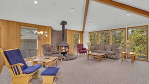 A cozy living room with a fireplace, wooden furniture, and large windows offering a view of the outdoors. - Unique, spacious, private deck, HT, great room (Sunriver)