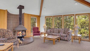 A spacious living room with a fireplace, wooden beams, large windows, and a red armchair. - Unique, spacious, private deck, HT, great room (Sunriver)