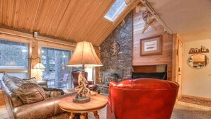 Ein gemütliches Wohnzimmer mit Kamin aus Stein, Ledermöbeln und einem großen Fenster mit Blick auf einen Wald.