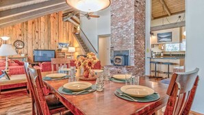 Zona pranzo con tavolo in legno apparecchiato per quattro persone, camino in mattoni e cucina sullo sfondo.