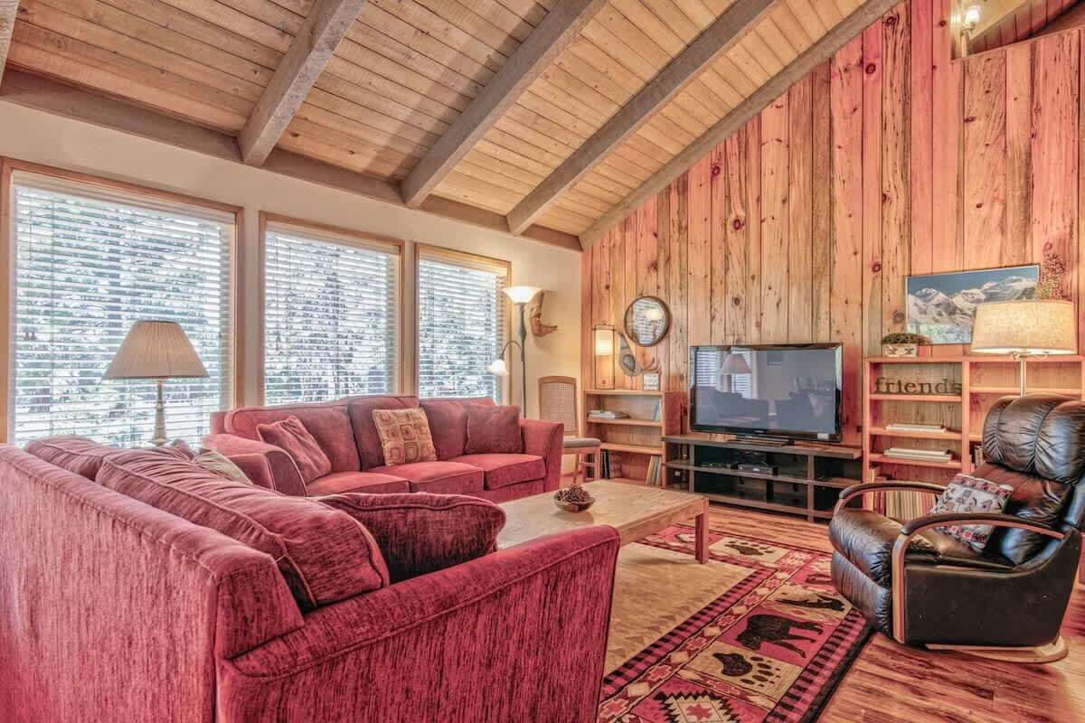 A cozy living room with a red sofa, a leather armchair, and a television. There are wooden walls and a large window with blinds.