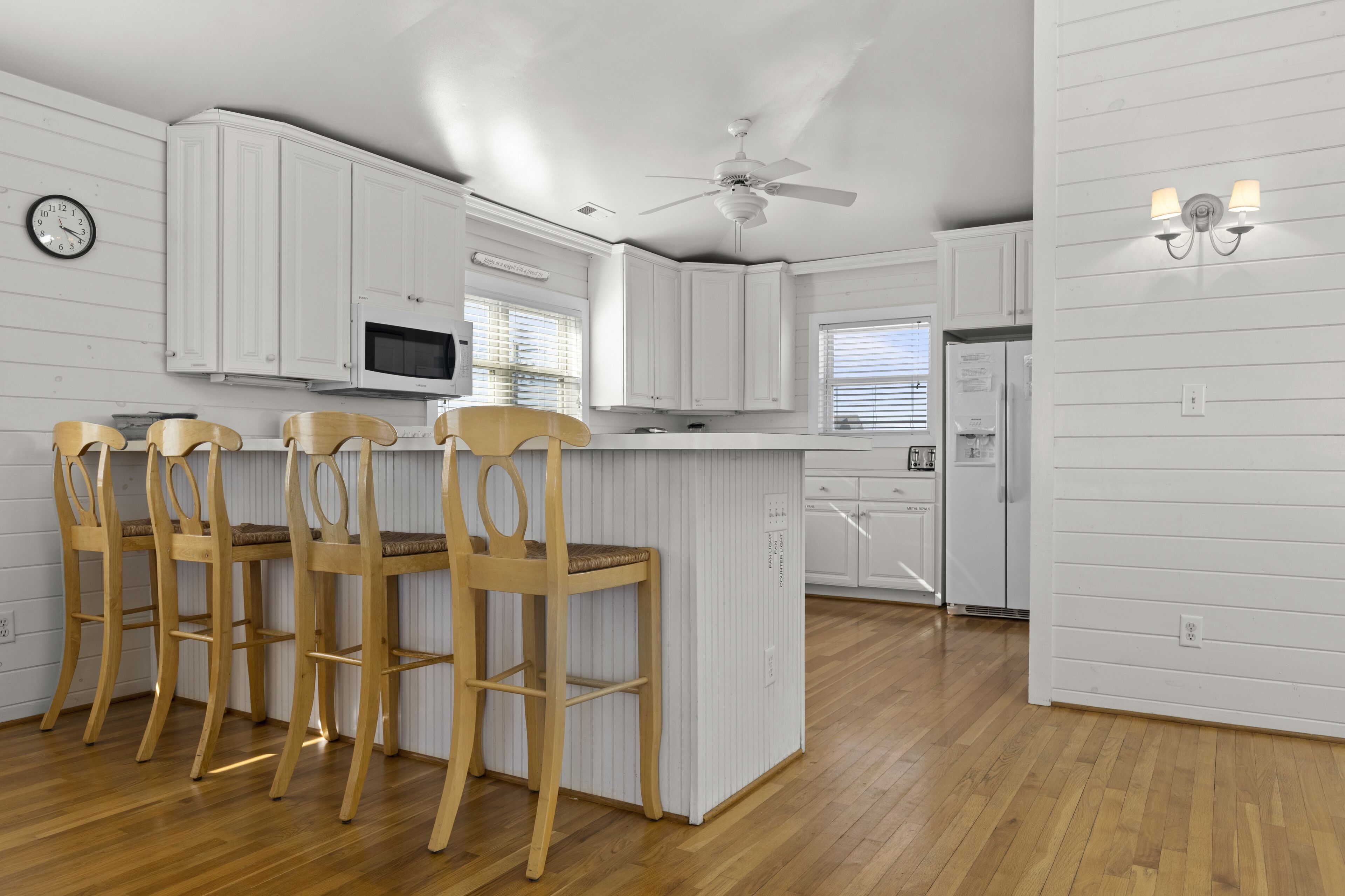Una cocina con armarios blancos, una isla de madera con taburetes de bar, un ventilador de techo y un reloj en la pared.