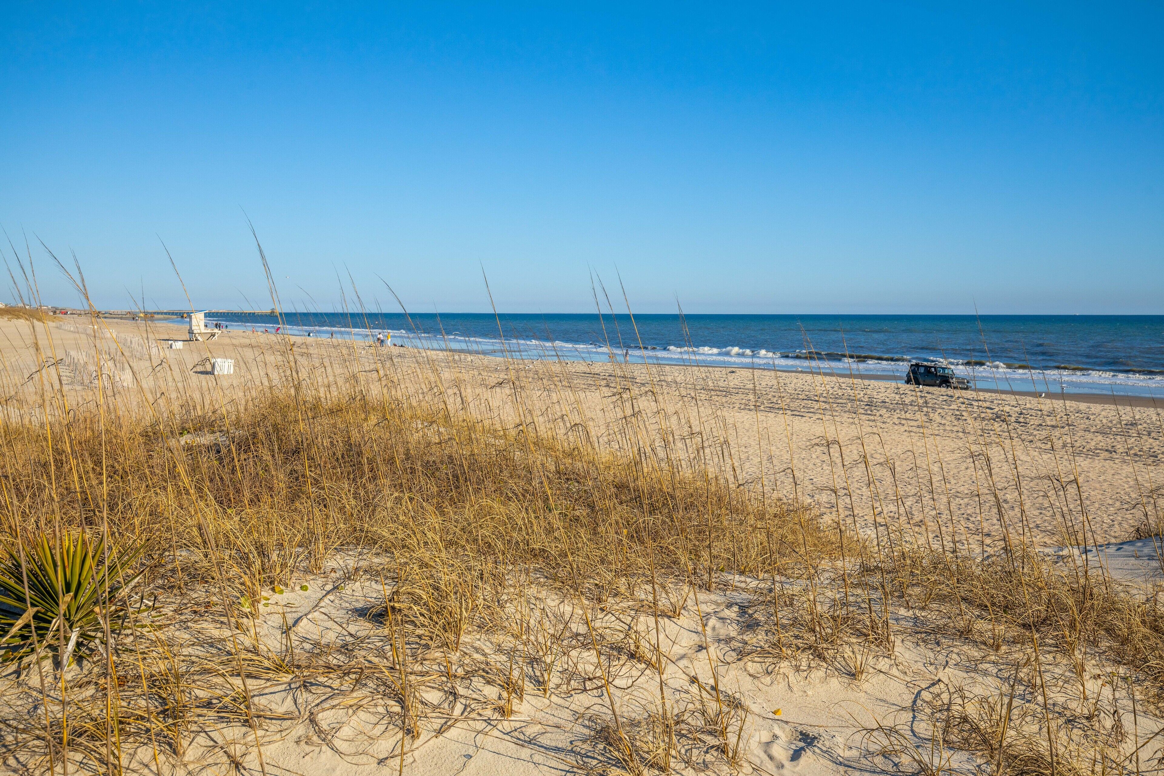 Une plage avec des dunes de sable, une végétation clairsemée et un poste de surveillance des sauveteurs au loin.