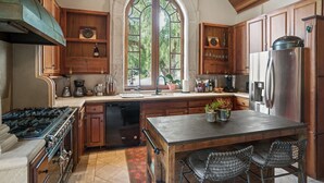 A kitchen with wooden cabinets, a large island with bar stools, a stainless steel refrigerator, and a window with a view of trees.
