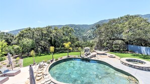 A swimming pool with a stone border, surrounded by a patio area with seating and umbrellas, set against a backdrop of trees and hills.