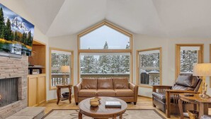 A cozy living room with a fireplace, a large window with a view of snow-covered trees, a brown leather sofa, and a wooden coffee table. - AC, pet friendly, inside Sunriver, EV charger (Sunriver)