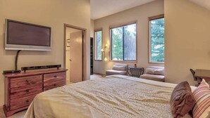 Un dormitorio con una cama grande, un televisor montado en la pared, un aparador y una ventana con vistas a árboles.