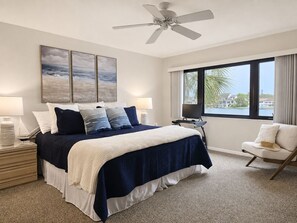 A bedroom with a bed, bedside tables, a chair, a ceiling fan, a window with a view, and artwork on the wall.