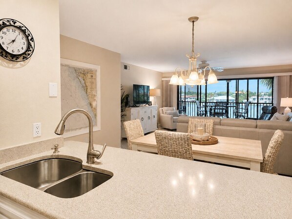 A kitchen with a double sink, a dining area with wicker chairs, a sofa, and a balcony with a view.