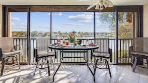 A balcony with a table set for two, chairs, a ceiling fan, and a view of a body of water and buildings. - AJ's Bayside Treasure - Outstanding Waterfront Views & Private Beach Access! (Sarasota)
