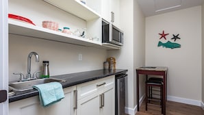 A kitchen with white cabinets, a black countertop, a sink, a microwave, and a small wooden table with a stool.
