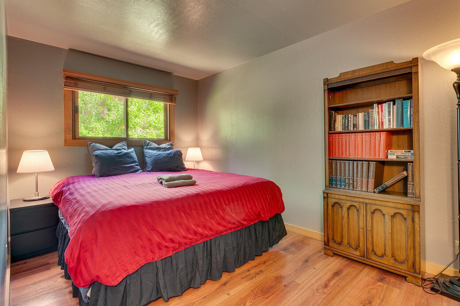 A bedroom with a large bed, a bookshelf, and a window with blinds.