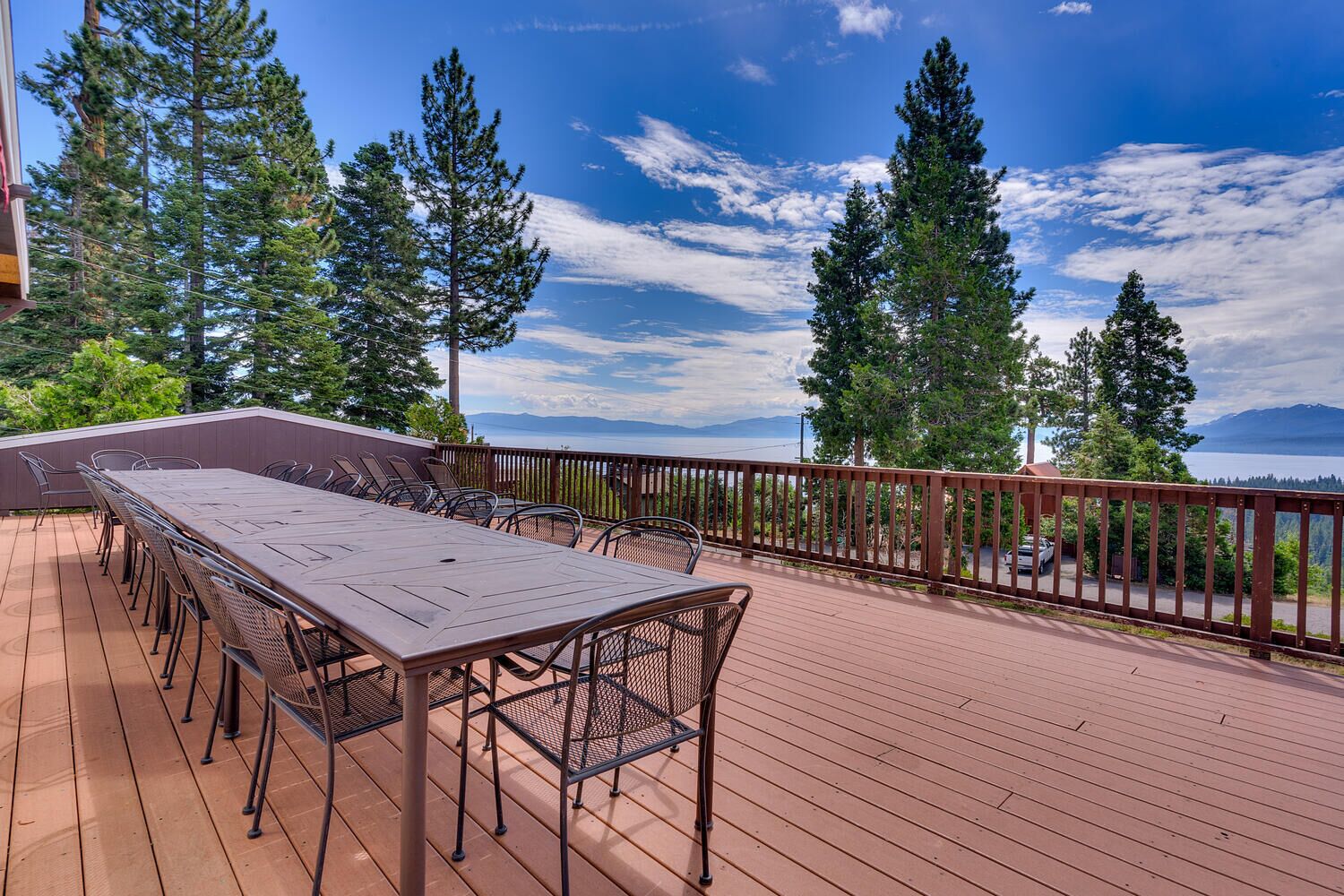 A wooden deck with a long table and chairs, surrounded by a railing, overlooking a body of water and mountains.