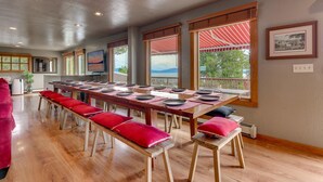 A spacious dining area with a long wooden table set for multiple people, benches with red cushions, and large windows offering a view of the outdoors.