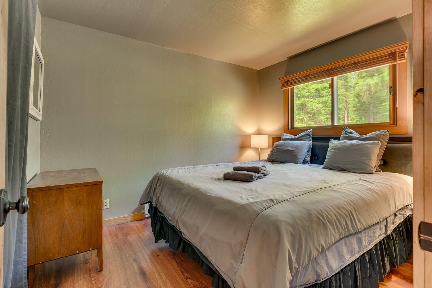 A bedroom with a bed, a nightstand, a window with a view of trees, and a lamp.