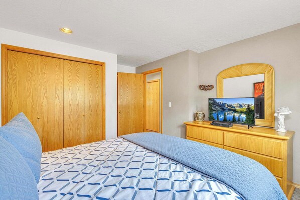 Un dormitorio con cama, armario de madera, televisor sobre un aparador de madera y espejo en la pared.