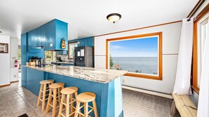 Une cuisine avec un îlot central bleu, des tabourets en bois, une vue sur la mer et une grande fenêtre.