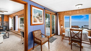 Une pièce chaleureuse avec une cheminée, des murs en bois, un coin repas avec une table et des chaises, et de grandes fenêtres offrant une vue sur la mer.