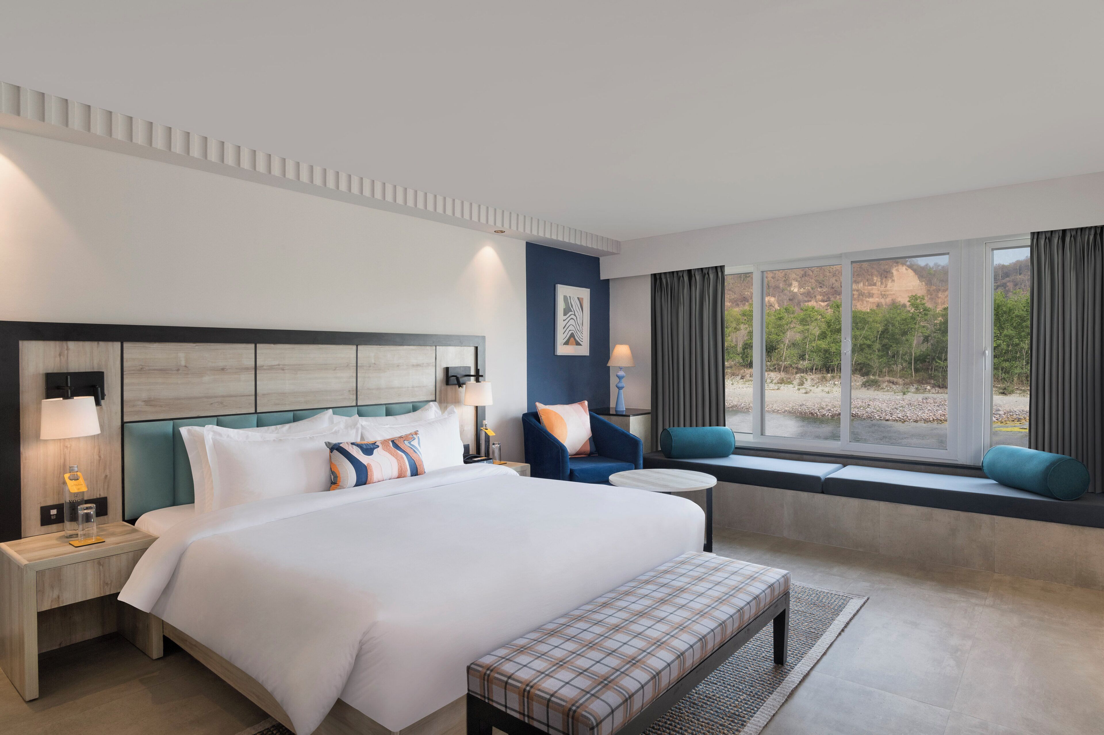 Habitación de hotel con una cama grande, un banco, una ventana con cortinas y vistas al exterior.