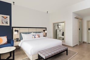 A hotel room with a bed, bedside table, chair, and a bathroom with a shower and toilet. - voco Jim Corbett by IHG (Ramnagar)