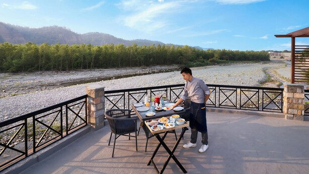 Een terras met een gedekte tafel, omringd door stoelen, met zicht op een rivier en beboste heuvels.