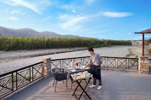 Un área de comedor al aire libre con una mesa dispuesta para una comida, rodeada de sillas y vista a un río y colinas boscosas al fondo.
