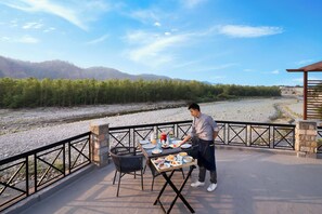 An outdoor dining area with a table set for a meal, surrounded by chairs, and a view of a river and forested hills in the background. - voco Jim Corbett by IHG (Ramnagar)